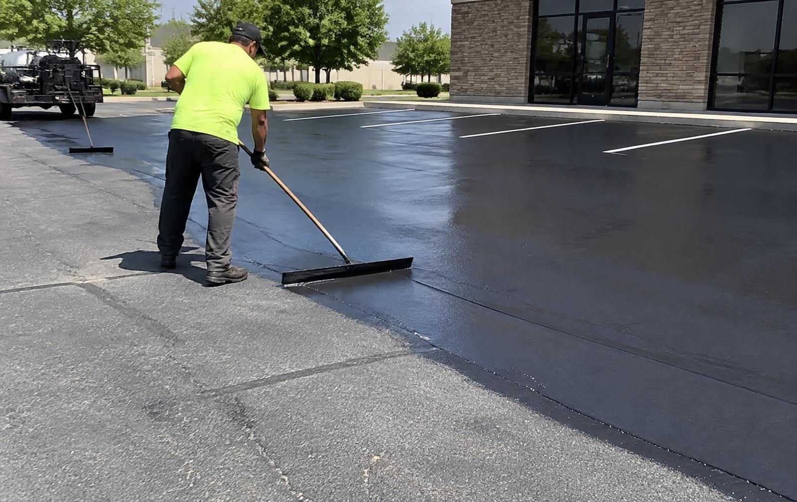Worker applying black asphalt sealcoat with a squeegee across a commercial parking lot